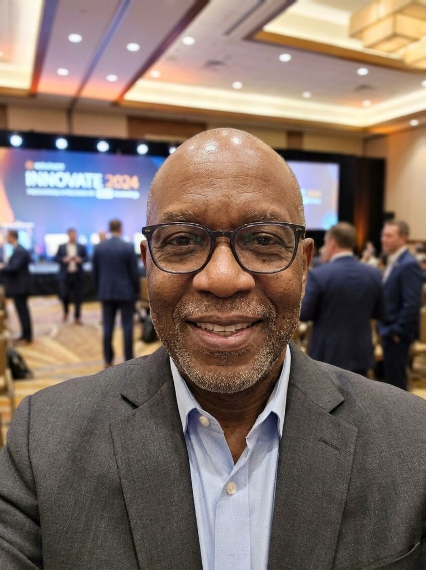 A smiling man in glasses and a grey blazer at a corporate conference in a hotel ballroom, with an INNOVATE 2024 stage screen and delegates behind him