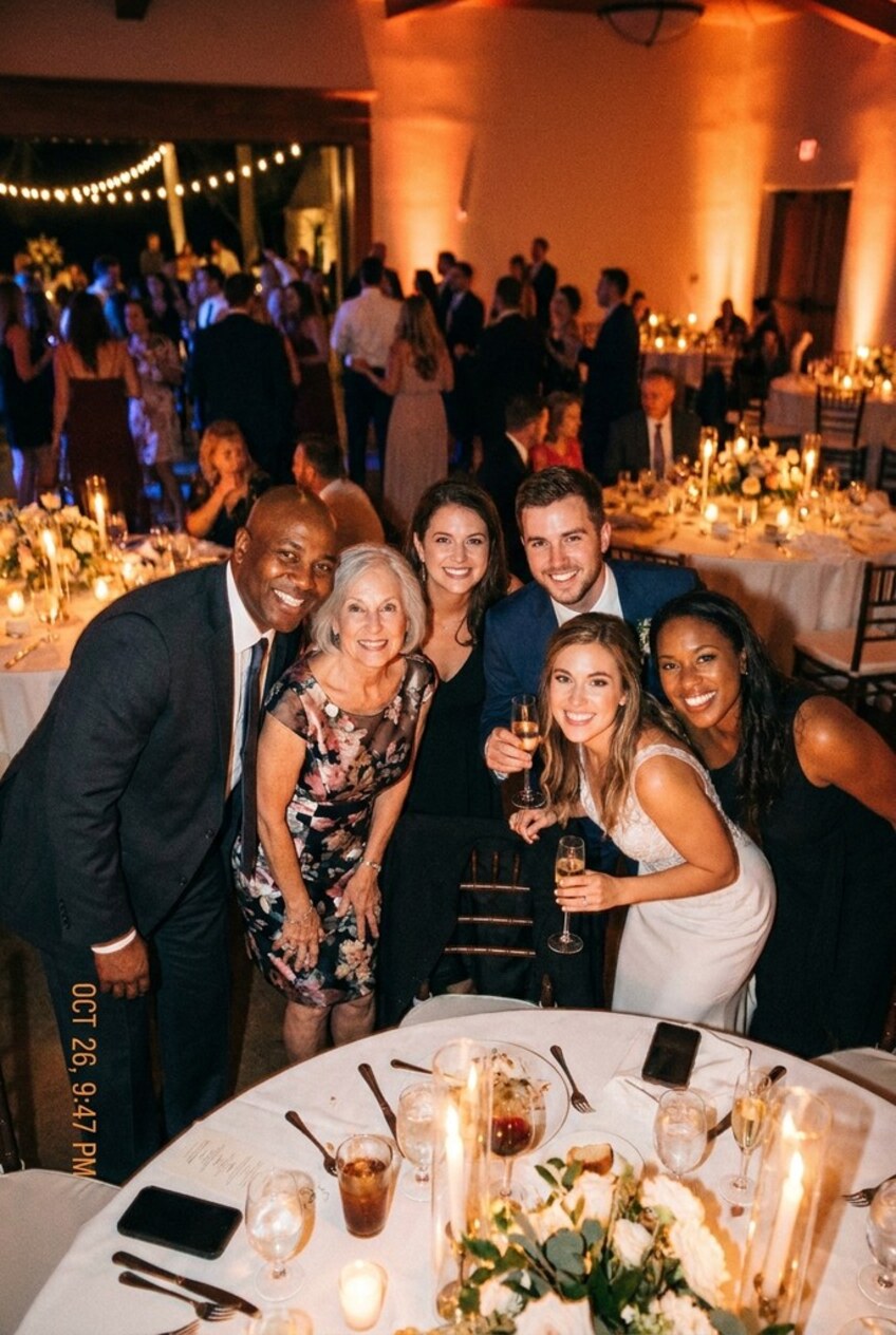 Six guests leaning in around a round banquet table at the reception, the bride in white lace among them