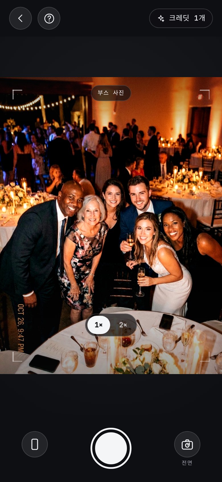 The AI photobooth in the app: a camera of its own, open on a group of guests leaning in at a candlelit reception, with a “booth shot” badge across the viewfinder, a help button top-left and the booth credits left top-right